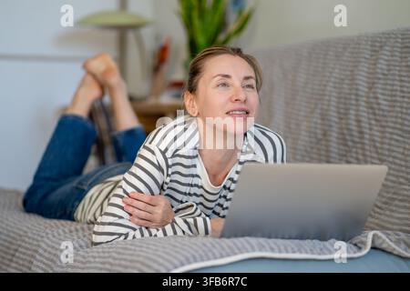 Donna positiva e pensierosa sdraiata sul divano, naviga online sul laptop, rilassandosi a casa nel fine settimana. Foto Stock