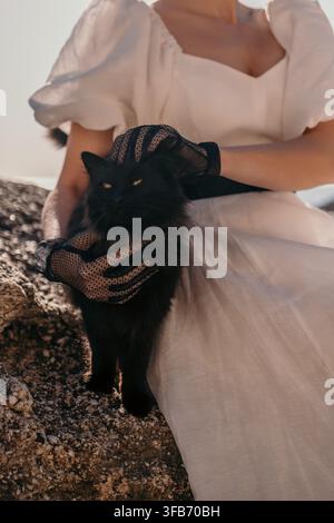Scena da spiaggia di una donna gatta, una persona in abito di lino bianco e guanti di pizzo che tiene un felino nero su scogli costieri Foto Stock