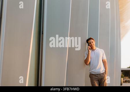 Un giovane uomo si appoggia rilassato contro un'architettura elegante, sorridendo mentre parla al telefono. Il tramonto proietta una luce calda intorno a lui. Foto Stock