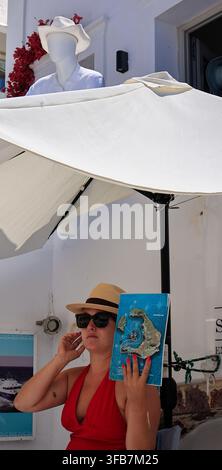 Oia, Santorini, Grecia - 4 agosto 2026: Una turista con un cappello di paglia tiene una mappa dell'isola, riecheggiando un cappello corrispondente su un manichino del negozio dietro di lei Foto Stock
