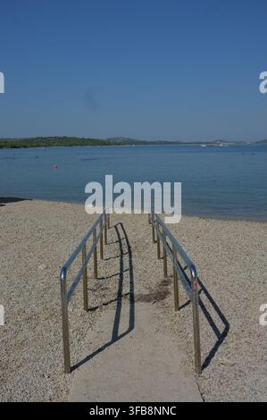 Ringhiere metalliche guidano giù per una riva di ciottoli verso acqua blu calma, con piccole boe, basse colline verdi e costa lontana sotto un vasto cielo sereno, creano Foto Stock