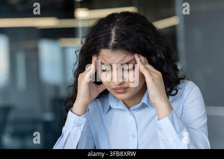 Giovane donna d'affari con capelli ricci scuri e camicia azzurra che sente un forte mal di testa o emicrania, preme le mani sulle tempie, soffre di dolore, stress e burnout in un ambiente d'ufficio moderno Foto Stock