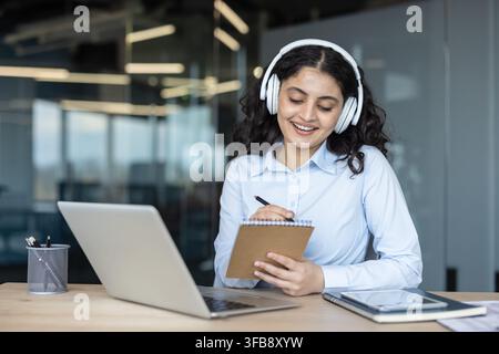 Giovane donna sorridente, con le cuffie e che scrive su un quaderno, prende appunti durante una lezione online o una riunione di lavoro utilizzando un laptop in un ambiente di lavoro contemporaneo Foto Stock
