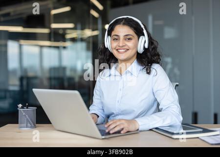 Giovane donna che guarda la telecamera, sorridendo mentre indossa cuffie e digita su un laptop, impegnata nel supporto clienti o nella comunicazione online in un ufficio moderno Foto Stock