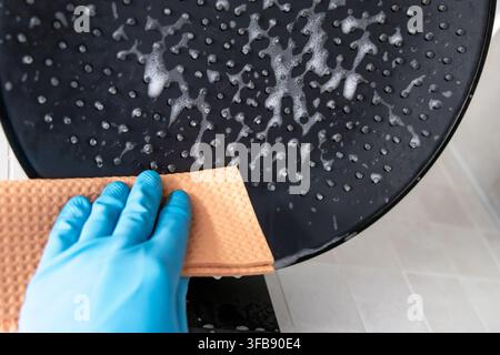 Prima piano di una mano che indossa un guanto protettivo blu che strofina un soffione nero con una spugna arancione ruvida e schiuma di sapone Foto Stock