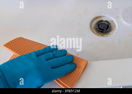 La mano di una persona con un guanto protettivo blu strofina la superficie sporca della vasca da bagno con una spugna arancione, concentrandosi sulla sporcizia intorno all'area di scarico Foto Stock
