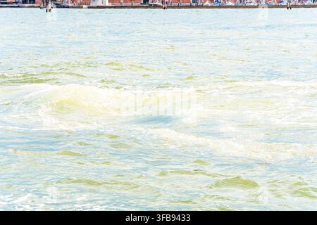 Increspature d'acqua illuminate dal sole e onde schiumose riempiono il primo piano vicino a una lontana passeggiata sul lungomare. Foto Stock