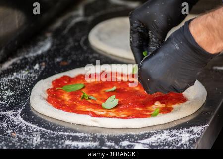 Pizzaiolo con guanti neri che prepara una pizza allo stile napoletano su un bancone di metallo infarinato in una pizzeria Foto Stock