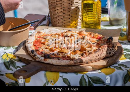 Pizza cotta nel forno a legna in stile napoletano appena sfornata con funghi selvatici, servita su una tavola di legno sopra una tovaglia a motivi di limone Foto Stock
