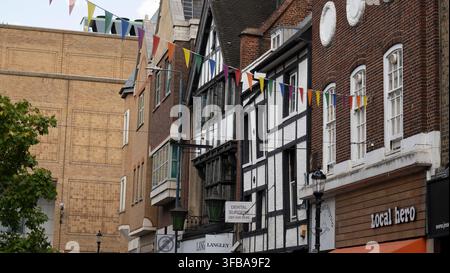 Un'inquadratura urbana a bassa angolazione che mette in risalto le facciate storiche in stile Tudor con struttura a graticcio accanto alle vetrine dei negozi in mattoni rossi su High Street a Kingston upon Thames, Foto Stock
