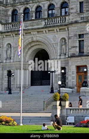 Una vista ravvicinata dell'arco d'ingresso in pietra degli edifici del Parlamento della British Columbia con un padre e un bambino seduti sul prato. Foto Stock