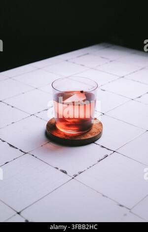 Cocktail rosso con un cubetto di ghiaccio trasparente in un bicchiere da roccia su un sottobicchiere di legno su piastrelle bianche, fotografia minimalista d'atmosfera delle bevande Foto Stock