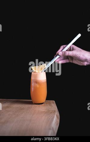 Mano di barista che usa una pinza per guarnire un cocktail all'arancia con una fetta di arancia su un tavolo di legno su uno sfondo nero Foto Stock