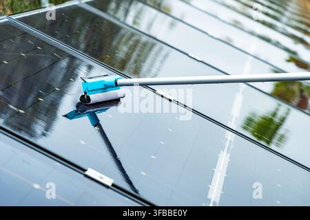 Una vista ravvicinata mostra uno strumento a spugna che pulisce una fila di pannelli solari fotovoltaici, riflettendo l'ambiente circostante Foto Stock