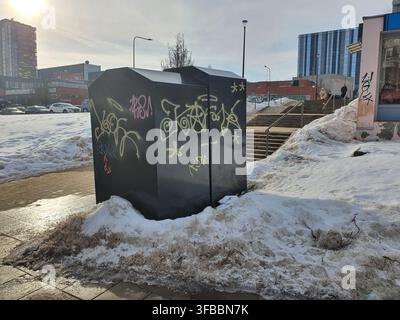 Due grandi cassonetti neri o bidoni per i rifiuti coperti da colorati graffiti si ergono in modo prominente su un marciapiede urbano parzialmente coperto di neve. Il Foto Stock
