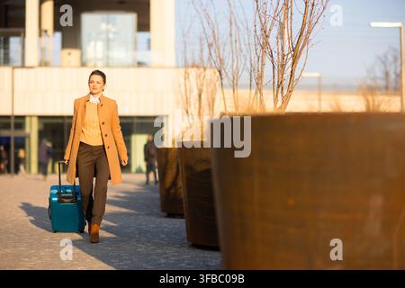 Una donna elegante in un cappotto marrone chiaro e maglione giallo avanza con una valigia rotante blu, riflettendo lo stile di vita moderno di un viaggiatore connesso i Foto Stock