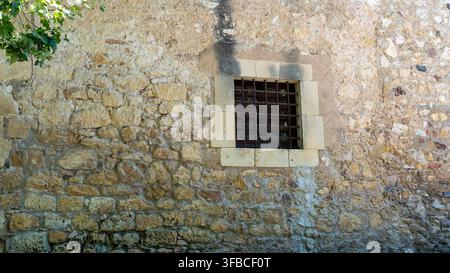 Primo piano di un vecchio muro di pietra rustico con una piccola finestra sbarrata, muratura consumata e pietra naturale texturizzata su un edificio storico Foto Stock
