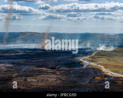 Diavoli di polvere e vortici di fumo che si alzano sopra una brughiera carbonizzata, ampia cicatrice di incendio ancora fumante lungo una strada di campagna sui Llangynidr Moors nel South Wa Foto Stock