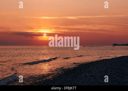 Trascinante tramonto vivace che proietta tonalità calde di arancione e rosa sull'acqua tranquilla del mare e sulle onde gentili che si infrangono su una riva di ciottoli, Dionisiou Bea Foto Stock