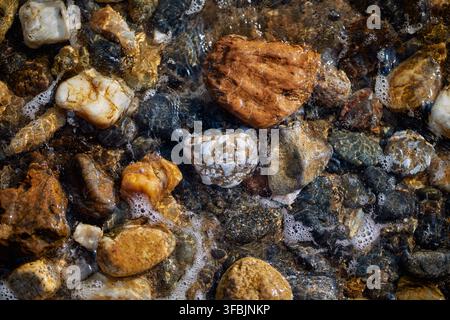 Prima piano ad angolazione alta di ciottoli bagnati colorati e schiuma bianca sul mare su una costa rocciosa in Grecia. Foto Stock