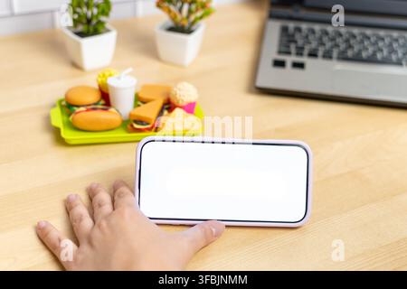 Mano che tiene uno smartphone orizzontale con schermo vuoto accanto a giocattoli di fast food su un tavolo di legno Foto Stock