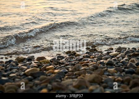 Prima piano di dolci onde marine che si infrangono su una raccolta di piccoli ciottoli e pietre lisci lungo una riva naturale, catturando il movimento tranquillo di Foto Stock