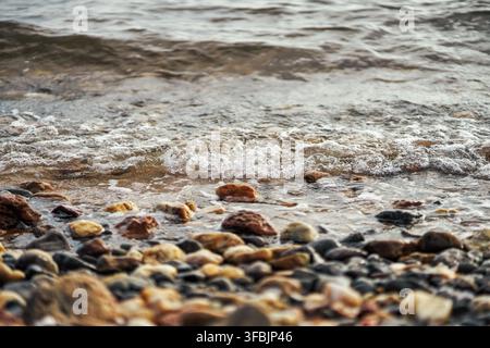 Acqua di mare limpida che si riversa su pietre e ciottoli colorati su una spiaggia naturale con schiuma che bolle dolcemente e motivi superficiali ondulati. Foto Stock