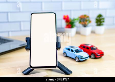 Smartphone con schermo vuoto su treppiede, macchinine giocattolo e laptop su una scrivania Foto Stock