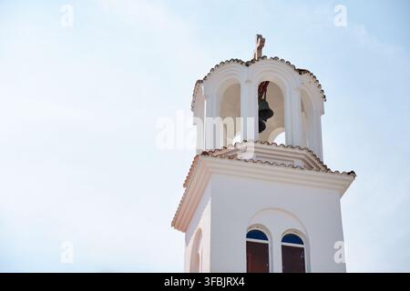 Vista ravvicinata di un campanile tradizionale bianco di chiesa ortodossa greca con una croce in cima, caratterizzato da aperture ad arco e campane appese all'interno sotto un Foto Stock