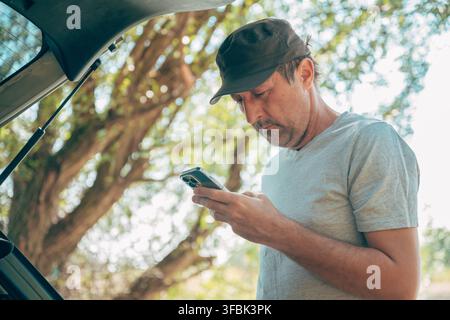 Conducente che controlla mappe di percorso online e messaggi su uno smartphone presso il portellone posteriore aperto del suo veicolo. Connettività moderna per viaggi, tecnologia di viaggio, digitale Foto Stock