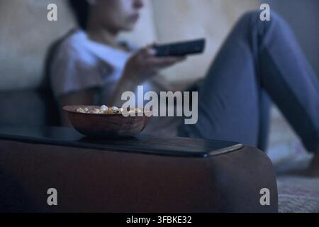 Vista ritagliata di una donna sfocata seduta sul divano con il telecomando mentre guarda la TV. Messa a fuoco selettiva su un piatto di popcorn. Concetto di insonnia, dipendenza, Foto Stock