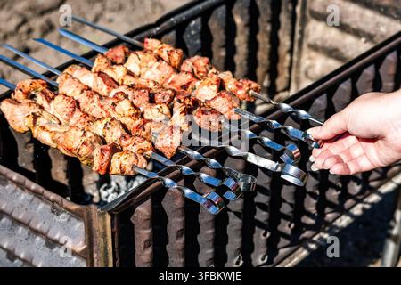 Primo piano della mano di una donna che si protende per toccare la maniglia metallica di uno spiedino di maiale succoso che arrostisce sul carbone all'aperto. Foto Stock