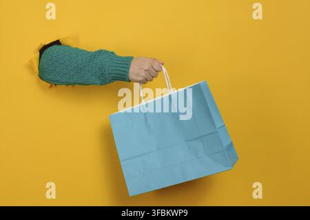 Una mano di donna, vestita con un maglione di lana verde, tiene un sacchetto di carta blu. La mano fuoriesce da un buco strappato in uno sfondo giallo brillante Foto Stock