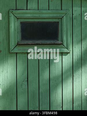 Un frammento di un vecchio muro di legno con una piccola finestra dipinta di verde Foto Stock