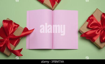 Un quaderno aperto con pagine rosa vuote, centrato, circondato da scatole regalo in carta kraft con archi rossi. Sfondo verde Foto Stock
