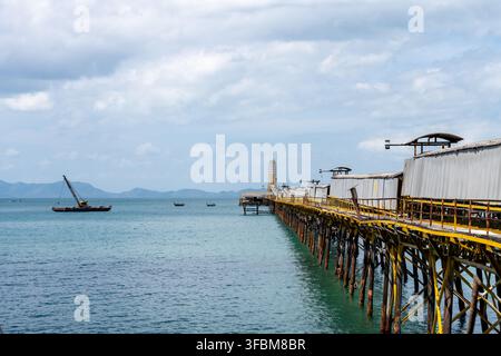 KRABI, THAILANDIA — 18 FEBBRAIO 2026: Un molo coperto per il trasporto di merci alla rinfusa si estende al largo vicino alla spiaggia di Tubkaek mentre una chiatta con gru galleggia accanto. Foto Stock