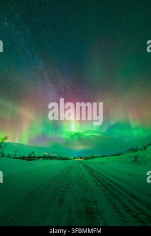 Vista delle vibranti aurore boreali che illuminano il cielo notturno sopra una strada innevata nel paesaggio artico di Tromso, Troms, Norvegia. Foto Stock
