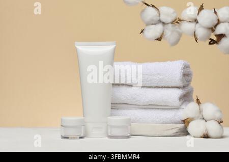 Pila di asciugamani bianchi, tubi e vasetti di plastica bianchi con cosmetici su sfondo beige Foto Stock