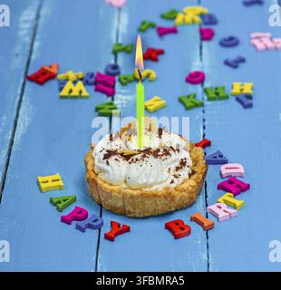 Dolce torta con crema bianca e un ardente cero giallo su sfondo blu sullo sfondo di legno, buon compleanno iscrizione Foto Stock