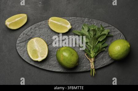 Lime intere e tagliate con foglie di menta fresche disposte su una superficie ruvida scura. Composizione perfetta per cibo, bevande o temi estivi Foto Stock
