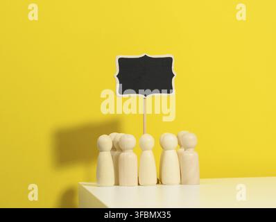 Gruppo di figurine di legno e un cartello vuoto su uno sfondo giallo. Il concetto di radunare persone per esprimere la loro volontà, dimostrazione, raduno e Foto Stock