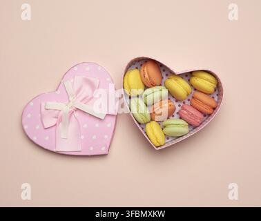 i macaron multicolore rotondi al forno si trovano in una scatola di cartone rosa a forma di cuore su uno sfondo beige, vista dall'alto Foto Stock