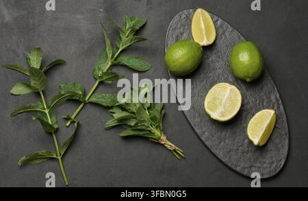 Lime intere e tagliate con foglie di menta fresche disposte su una superficie ruvida scura. Composizione perfetta per cibo, bevande o temi estivi Foto Stock