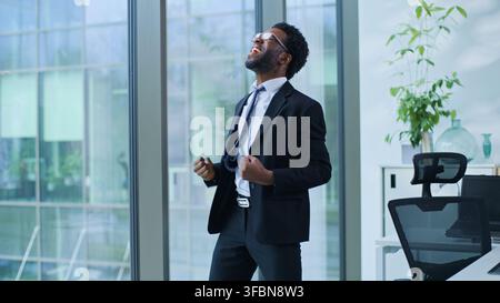 Uomo d'affari afroamericano felice che urla, vince, celebra, salta in ufficio, datore di lavoro maschile, uomo d'affari etnico, allegro, vittoria, celebrare, realizzazione Foto Stock