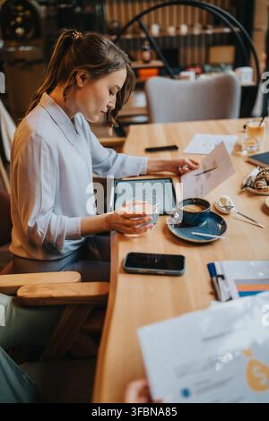 Donna d'affari che esamina documenti e un tablet mentre beve caffè durante un incontro informale in un bar Foto Stock