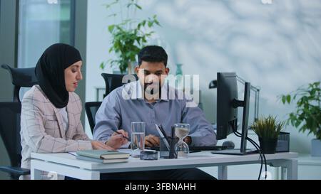 Due uomini d'affari indiani lavorano in squadra in ufficio con computer e documenti, sorridono, parlano, pianificano una strategia di progetto, colleghi arabi parlano del lavoro Foto Stock