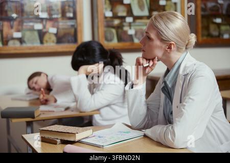 Studentessa di medicina giovane adulta caucasica che ascolta una lezione in aula mentre i compagni di classe dormono. L'immagine rappresenta il burnout accademico, perfetta per il marketing educativo Foto Stock