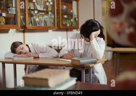 Adolescenti caucasiche e asiatiche in camici da laboratorio che studiano in aula di biologia, una dorme sulla scrivania. Concetto di affaticamento accademico, utile per il marketing educativo Foto Stock