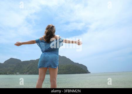 Vista posteriore di una giovane donna adulta che apre le braccia contemplando la meravigliosa isola di Phi Phi, Thailandia Foto Stock