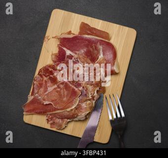 Pezzi sottili di jamon su un asse di legno, tavolo nero Foto Stock